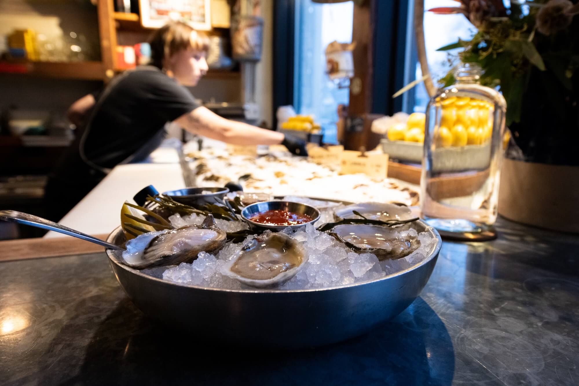 A chilled metal bowl filled with freshly shucked oysters sits on a bed of crushed ice, garnished with seaweed and accompanied by small metal cups containing cocktail sauce and mignonette. A long-handled spoon rests on the edge of the bowl. In the background, a person in a black shirt with short hair is reaching across a well-lit oyster bar, arranging or serving more oysters laid out on ice. The setting appears to be a cozy, bustling raw bar or seafood restaurant, with details like a galvanized metal bin of bright yellow lemons, floral arrangements, and soft natural light filtering in from a window, creating a welcoming and artisanal atmosphere.