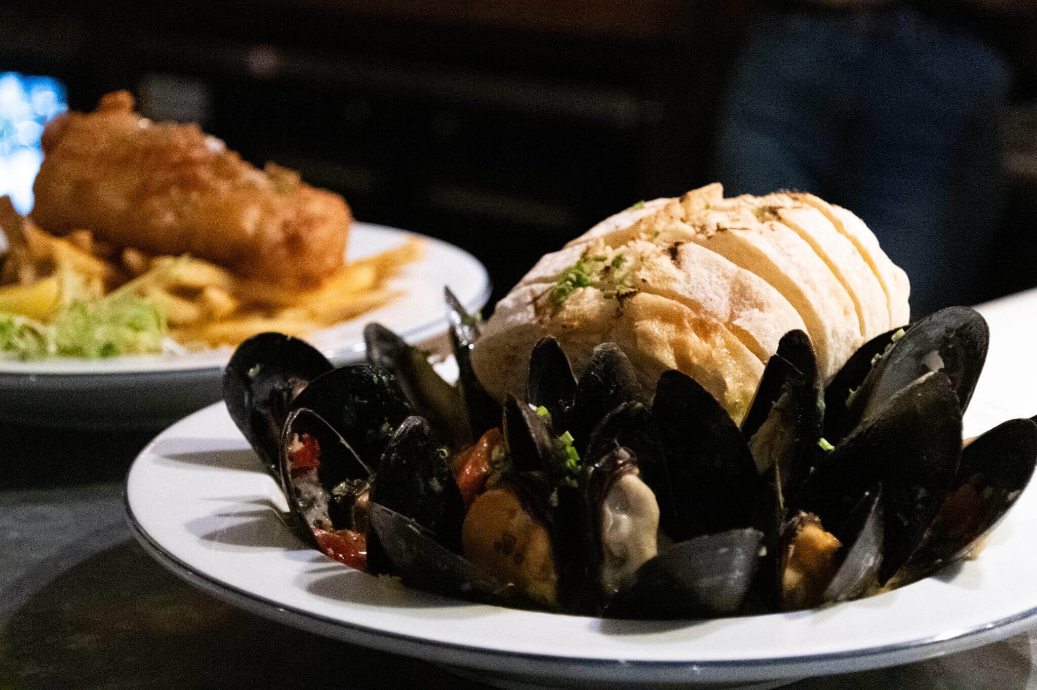 A close-up of a plated seafood dish featuring a generous serving of steamed mussels arranged in a circular pattern around the base of a thick, crusty sliced loaf of bread garnished with herbs. The mussels are interspersed with visible bits of tomato and green herbs. In the blurred background, a second plate can be seen holding a golden-brown battered fish fillet served atop a bed of French fries with a side of shredded lettuce, suggesting a classic fish and chips dish. The setting appears to be dimly lit, possibly a cozy restaurant or pub.